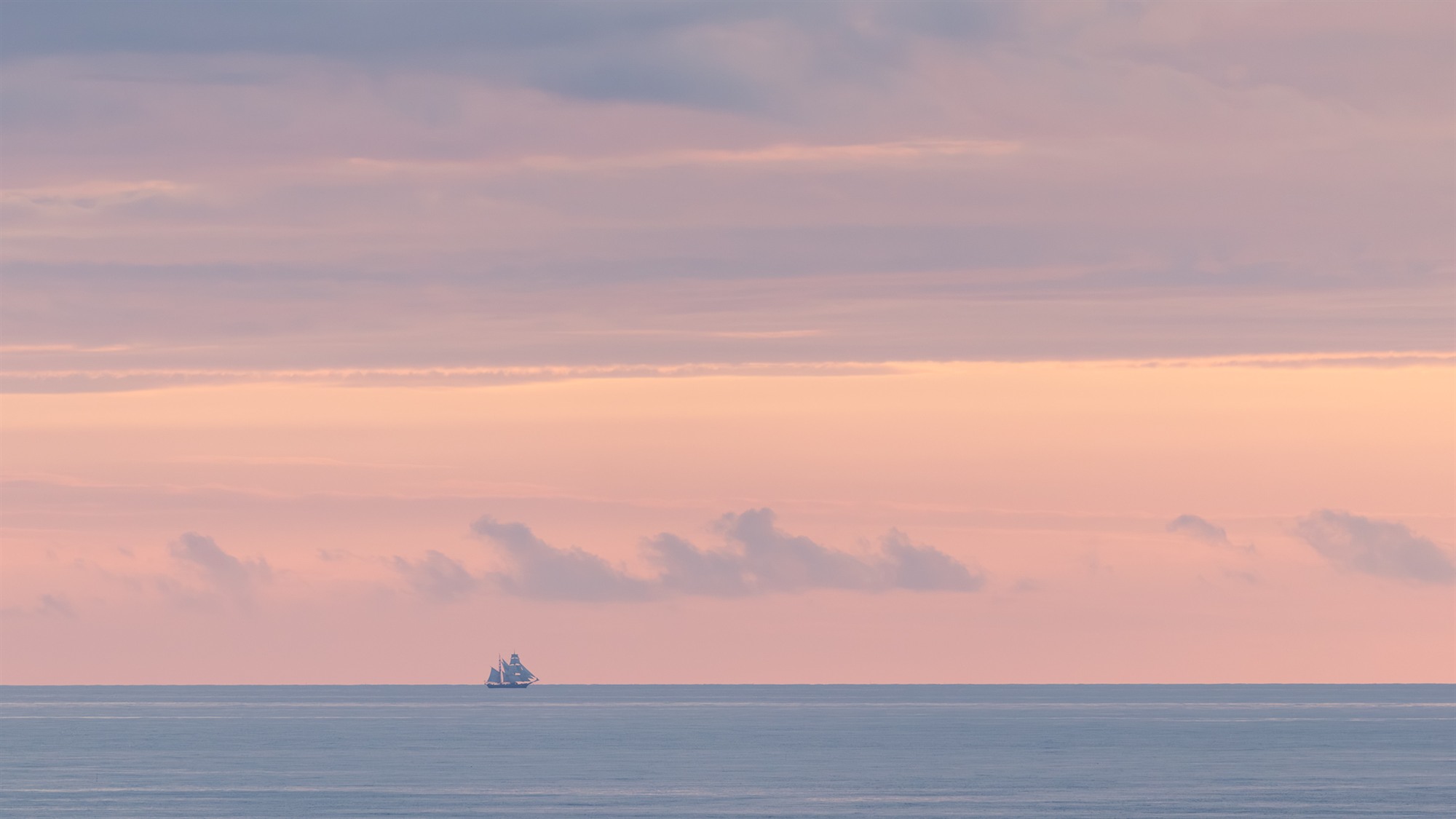 Ein kleines Schiff am Horizont unter einem pastellrosa und violett gefärbten Himmel.